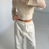 En person iført en hvid, åbenstrikket sweater samt hvide bukser står i et minimalistisk rum med lyse vægge og en hvid gulvflade.