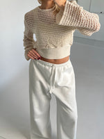 En person iført en hvid, åbenstrikket sweater samt hvide bukser står i et minimalistisk rum med lyse vægge og en hvid gulvflade.