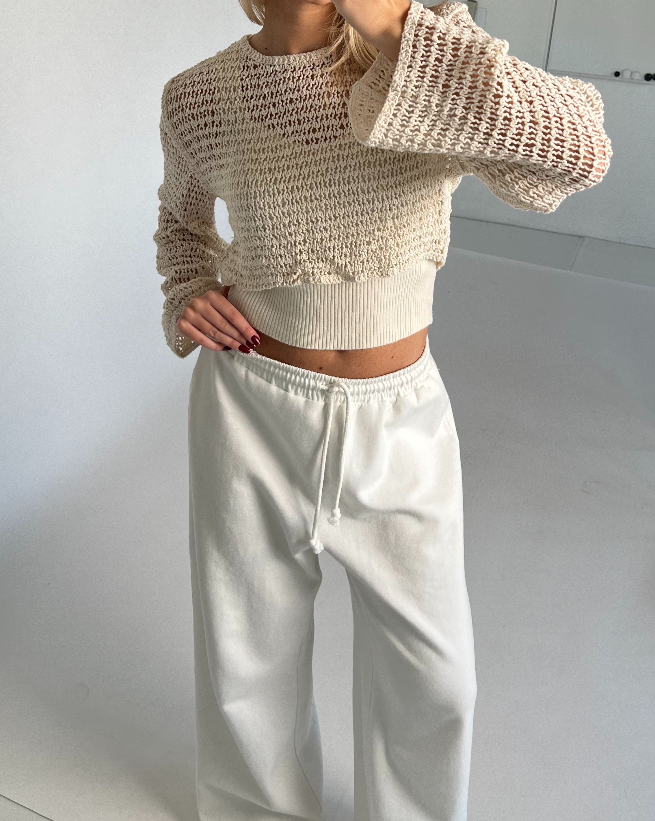 En person iført en hvid, åbenstrikket sweater samt hvide bukser står i et minimalistisk rum med lyse vægge og en hvid gulvflade.
