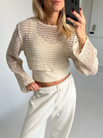 En person klædt i en cremefarvet, strikket trøje tager en selfie i et lyst rum. Personen bærer hvide joggingbukser og har en hånd på hoften.