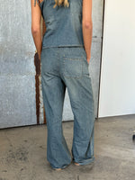 En person iført denim jumpsuit står med ryggen til, hænderne i lommerne. Baggrunden er en kombination af gråmetal og hvid væg på et betongulv.
