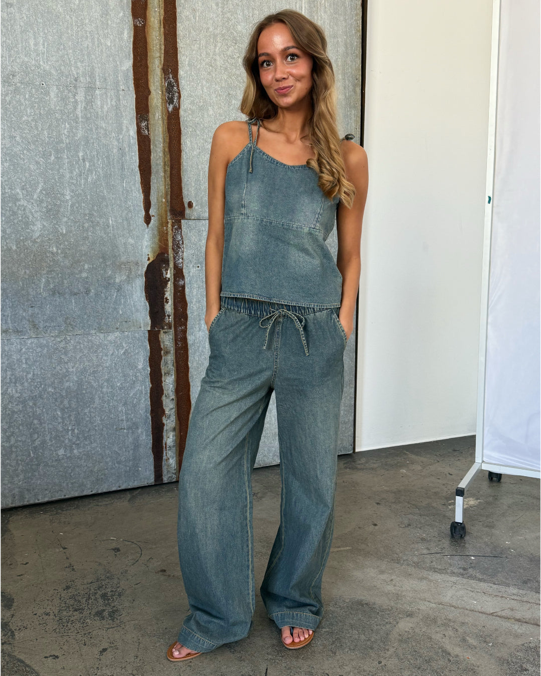 En kvinde i denim jumpsuit står afslappet med hænderne i lommerne. Hun smiler, omgivet af industrielle, rustikke vægge og betongulv.