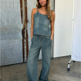 En kvinde i denim jumpsuit står afslappet med hænderne i lommerne. Hun smiler, omgivet af industrielle, rustikke vægge og betongulv.