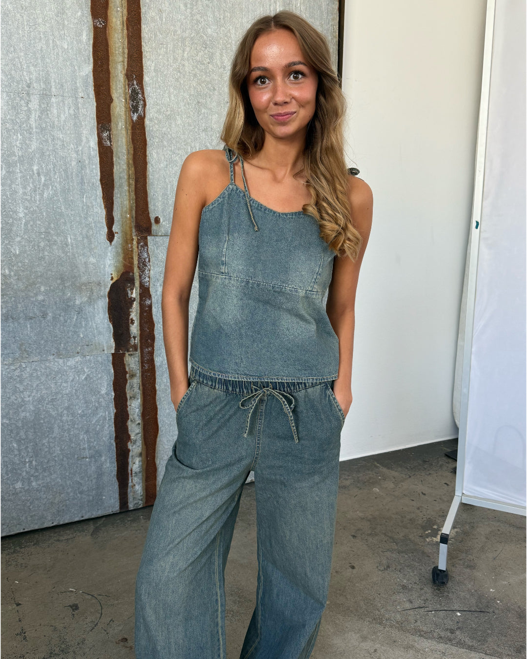 Kvinde i denim jumpsuit står afslappet med hænderne i lommerne, foran en rusten metalbaggrund og en hvid væg, med hård overgang i en fotostudiosetting.