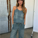 Kvinde i denim jumpsuit står afslappet med hænderne i lommerne, foran en rusten metalbaggrund og en hvid væg, med hård overgang i en fotostudiosetting.