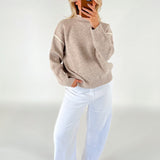 En person iført en beige sweater og hvide bukser står med hånden i lommen og tager et billede med en smartphone mod en neutral baggrund.