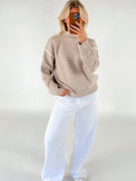 En person iført en beige sweater og hvide bukser står med hånden i lommen og tager et billede med en smartphone mod en neutral baggrund.