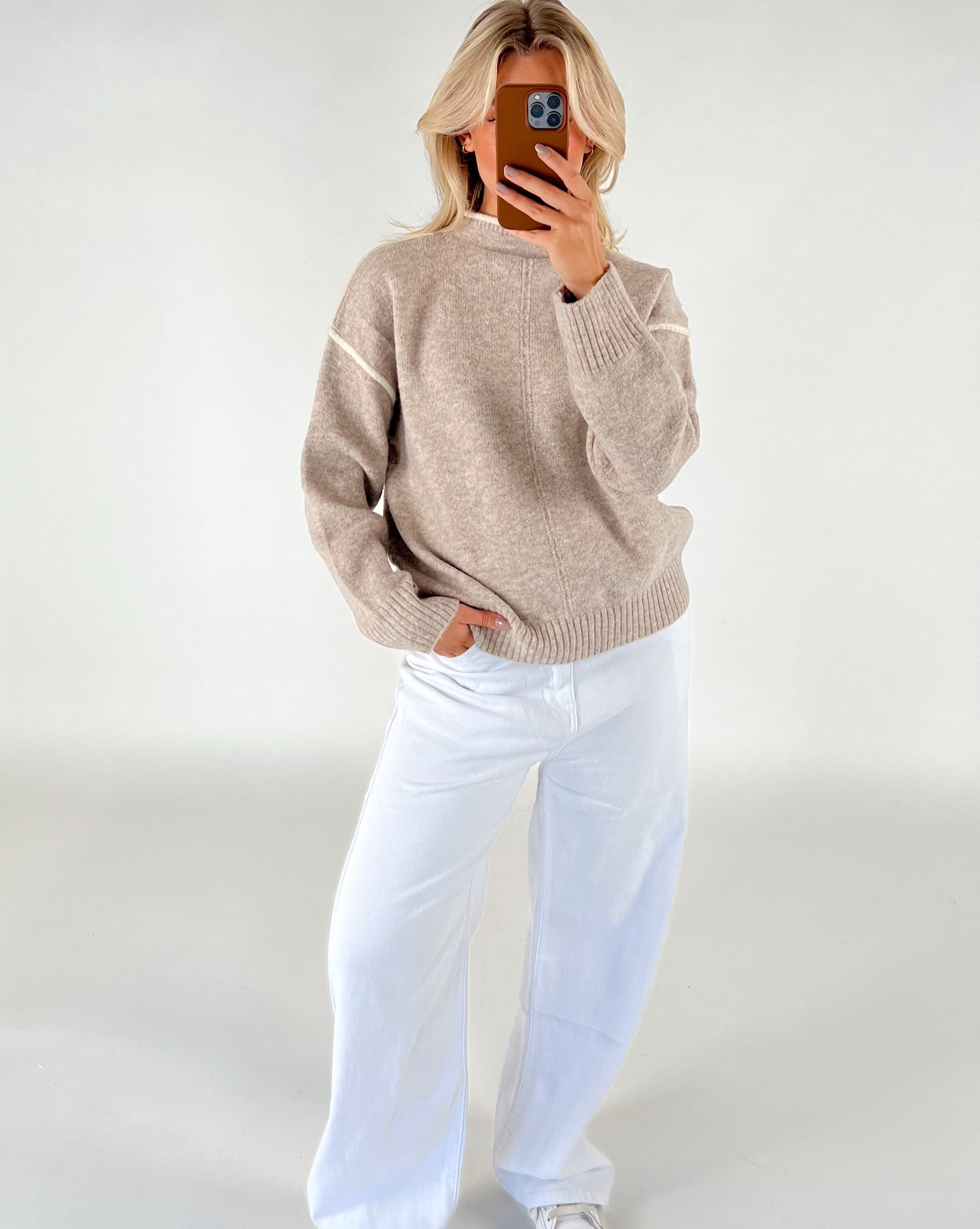 En person iført en beige sweater og hvide bukser står med hånden i lommen og tager et billede med en smartphone mod en neutral baggrund.