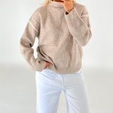 En person iført en beige sweater og hvide bukser tager et spejlbillede. Personen holder en mobiltelefon, og baggrunden er lys og ensfarvet.