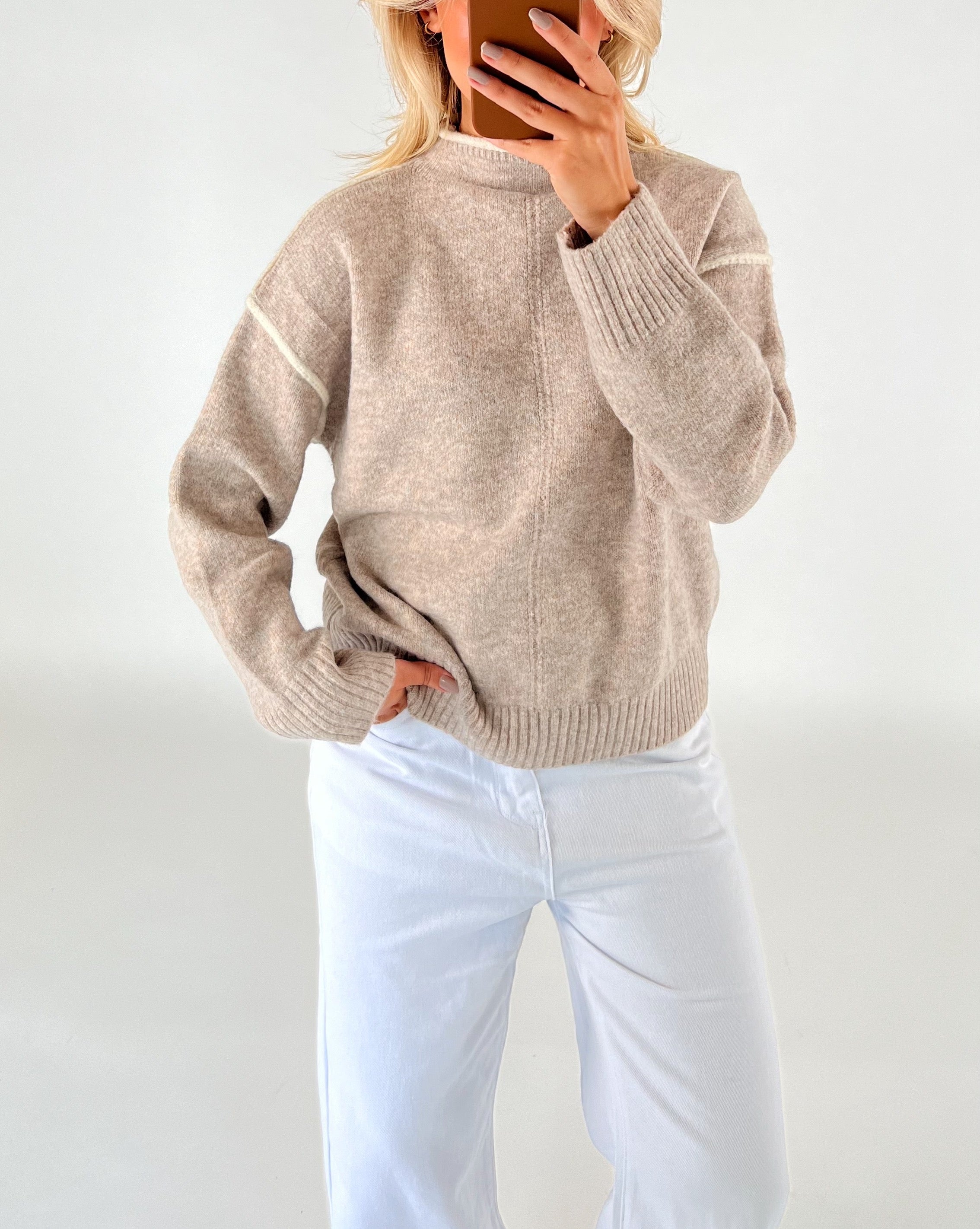 En person iført en beige sweater og hvide bukser tager et spejlbillede. Personen holder en mobiltelefon, og baggrunden er lys og ensfarvet.