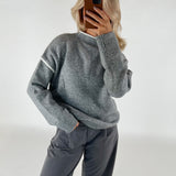 Person tager et billede af sig selv, iført en grå sweater og mørke bukser, står i et neutralt oplyst rum med hvid baggrund.