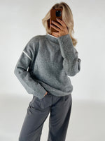 Person tager et billede af sig selv, iført en grå sweater og mørke bukser, står i et neutralt oplyst rum med hvid baggrund.