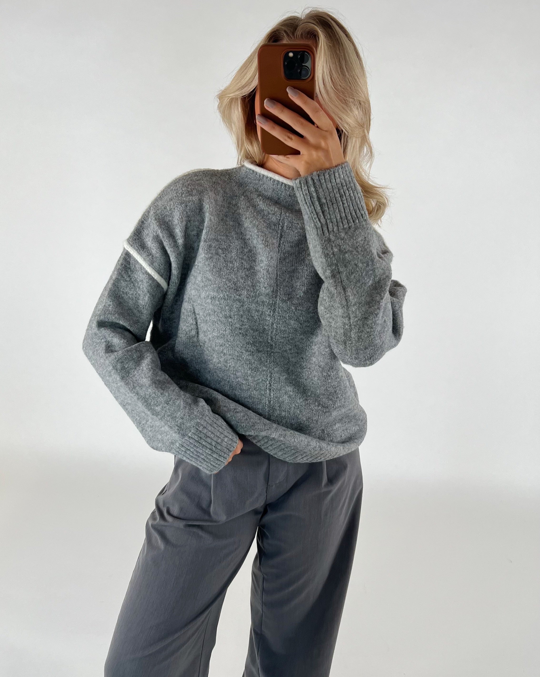 Person tager et billede af sig selv, iført en grå sweater og mørke bukser, står i et neutralt oplyst rum med hvid baggrund.