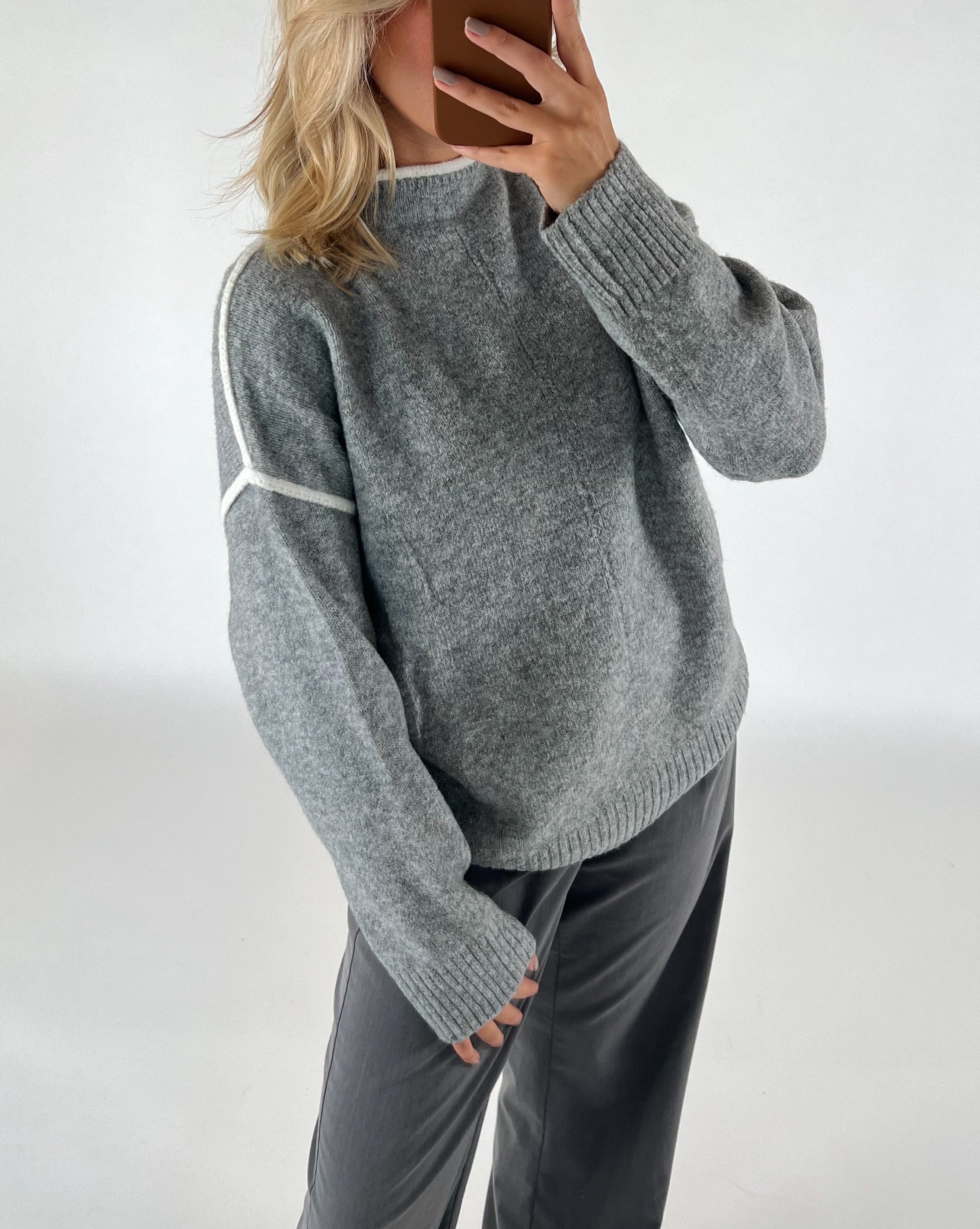 En person iført en grå sweater med hvide striber tager et spejlselfie med en smartphone, omgivende enkle og ensfarvede baggrund.
