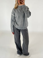 Person iført en løs, grå sweater og vide, mørkegrå bukser står i et minimalistisk rum, tager et spejlbillede med en smartphone.