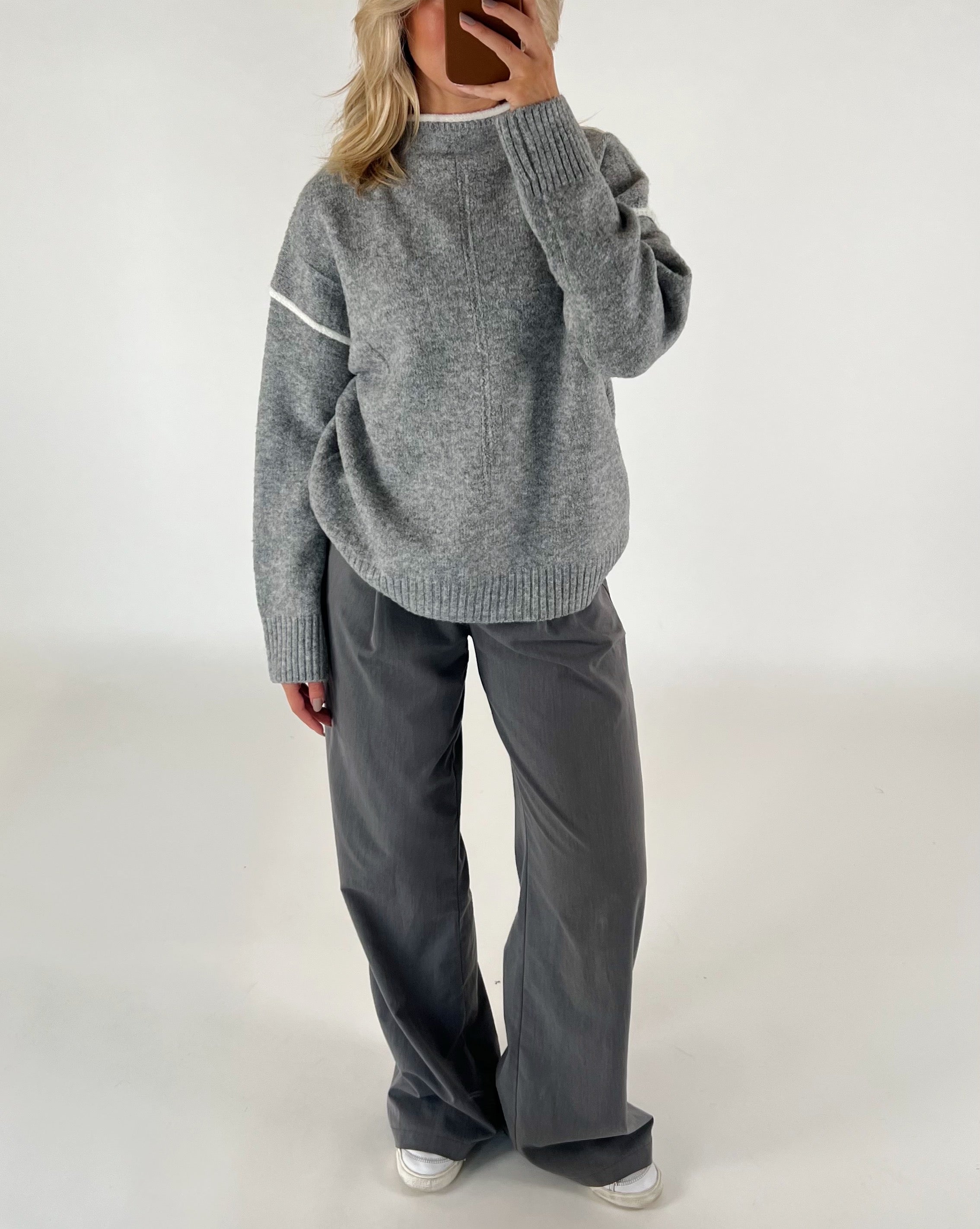 Person iført en løs, grå sweater og vide, mørkegrå bukser står i et minimalistisk rum, tager et spejlbillede med en smartphone.