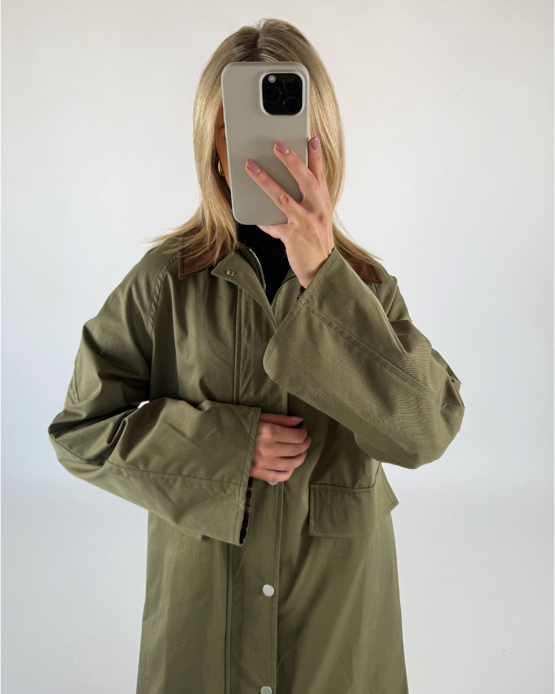 En person iført en olivengrøn frakke holder en smartphone foran ansigtet. Baggrunden er hvid og minimalistisk, hvilket fremhæver frakkens farve.