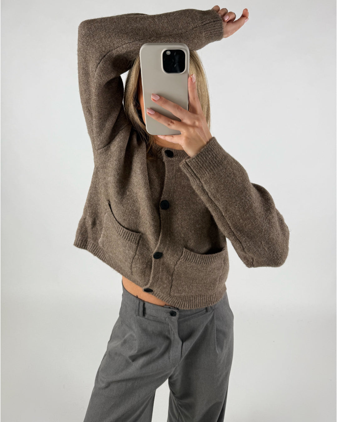 Person tager et spejlbillede med en smartphone iført en brun cardigan med sorte knapper. De står i et enkelt, hvidt rum og har løftet en arm op over hovedet.