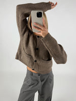 Person tager et spejlbillede med en smartphone iført en brun cardigan med sorte knapper. De står i et enkelt, hvidt rum og har løftet en arm op over hovedet.