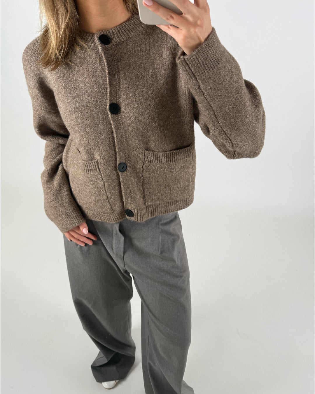 En person iført brun cardigan med lommer og sorte knapper tager et spejlbillede iført grå, vide bukser i et neutralt, lyst rum.