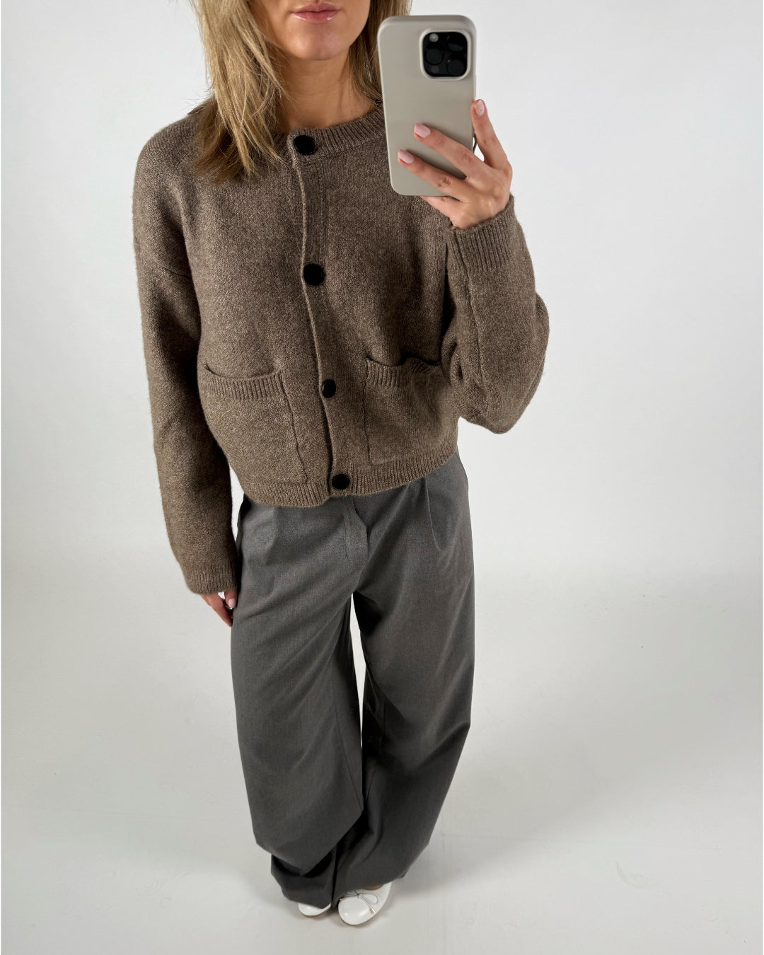 Kvinde holder en telefon og tager et spejlbillede. Hun har en brun cardigan og grå bukser på. Billedet tages i et neutralt, hvidt rum.
