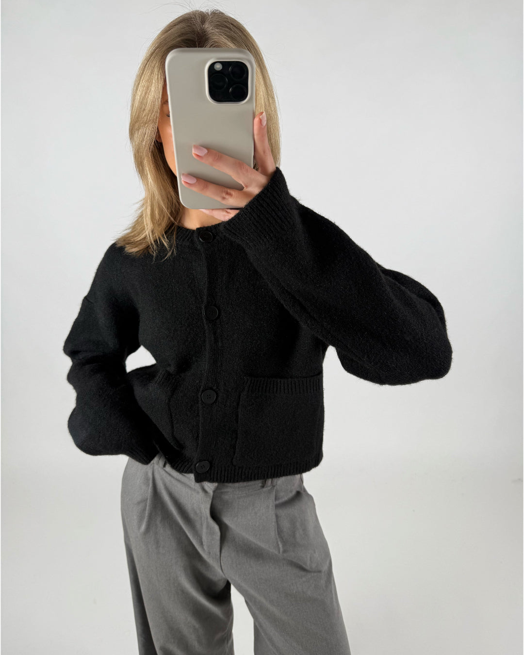 En person iført sort cardigan tager et spejlbillede med en smartphone. Personen har lyst hår, og bukserne er grå, i et neutralt rum.