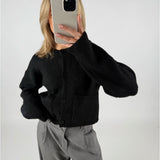 En person iført sort cardigan tager et spejlbillede med en smartphone. Personen har lyst hår, og bukserne er grå, i et neutralt rum.