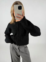 En person iført sort cardigan tager et spejlbillede med en smartphone. Personen har lyst hår, og bukserne er grå, i et neutralt rum.