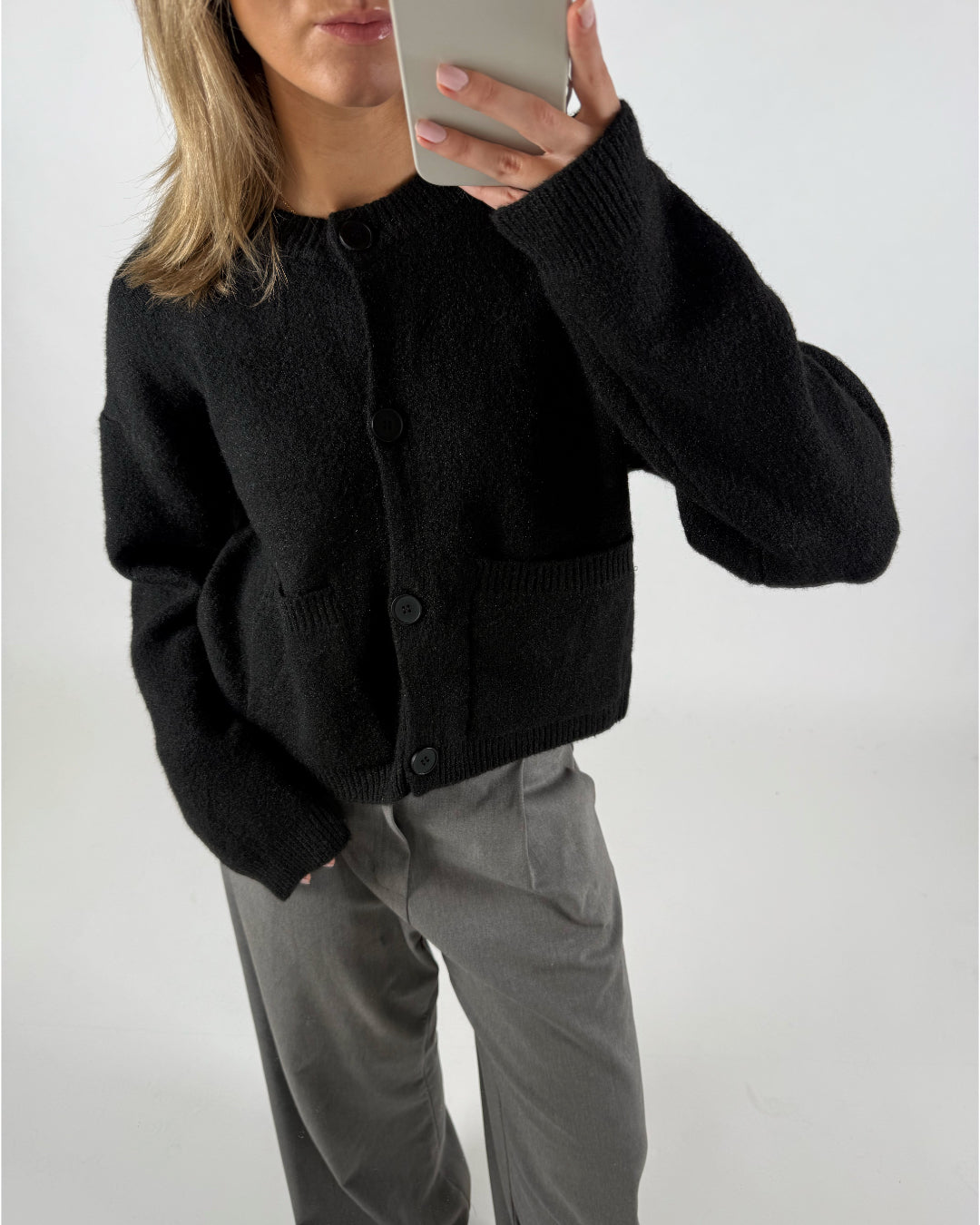 En person iført sort cardigan og grå bukser tager et spejlbillede med en smartphone i en lys baggrund.