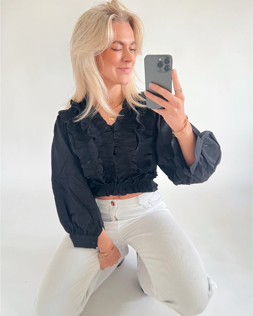 En person, iført en sort bluse og hvide bukser, tager en selfie med en smartphone. Personen knæler i et rum med en lys baggrund.