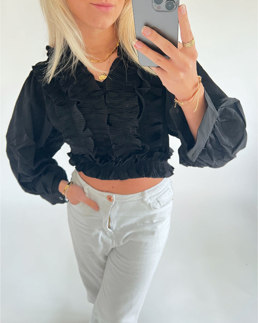 En person iført en sort bluse med flæser og hvide bukser tager et selfie med en mobiltelefon, mens de står foran en neutral baggrund. Personen har en halskæde og armbånd.