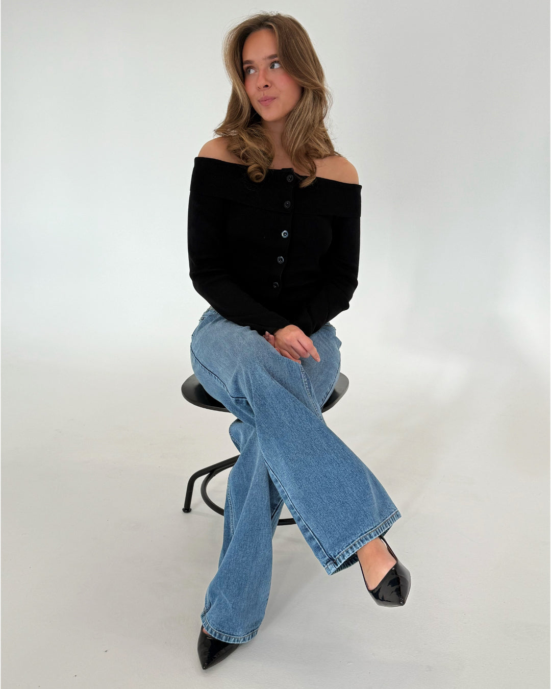 En kvinde sidder på en høj sort skammel. Hun bærer en sort off-shoulder bluse og blå jeans. Rummet omkring hende er hvidt og neutralt.