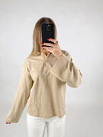 Person tager et billede med en smartphone; iført beige skjorte og hvide bukser. Personen er i et rum med en ensfarvet hvid baggrund.