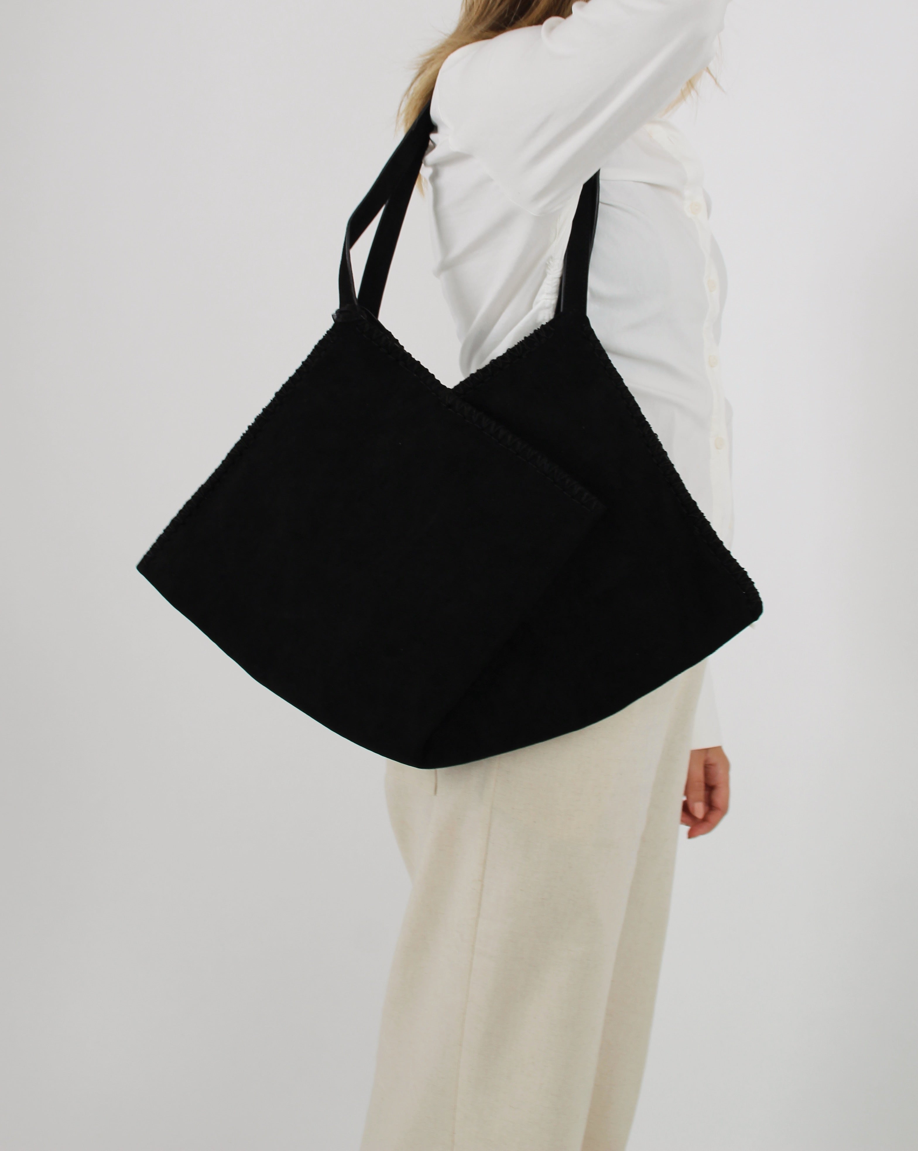 Sort taske bæres over skulderen af person i hvid skjorte og beige bukser; skaber en elegant og minimalistisk stil i neutral baggrund.