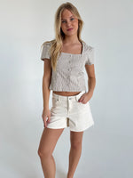 En kvinde står afslappet i en hvidblå stribet top og hvide shorts i et minimalistisk rum med lyse vægge.