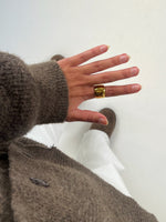 Hånd bærer en gylden ring og peger nedad. Personen har en brunt sweater og hvide bukser på. Støvler er synlige i baggrunden på en hvid baggrund.