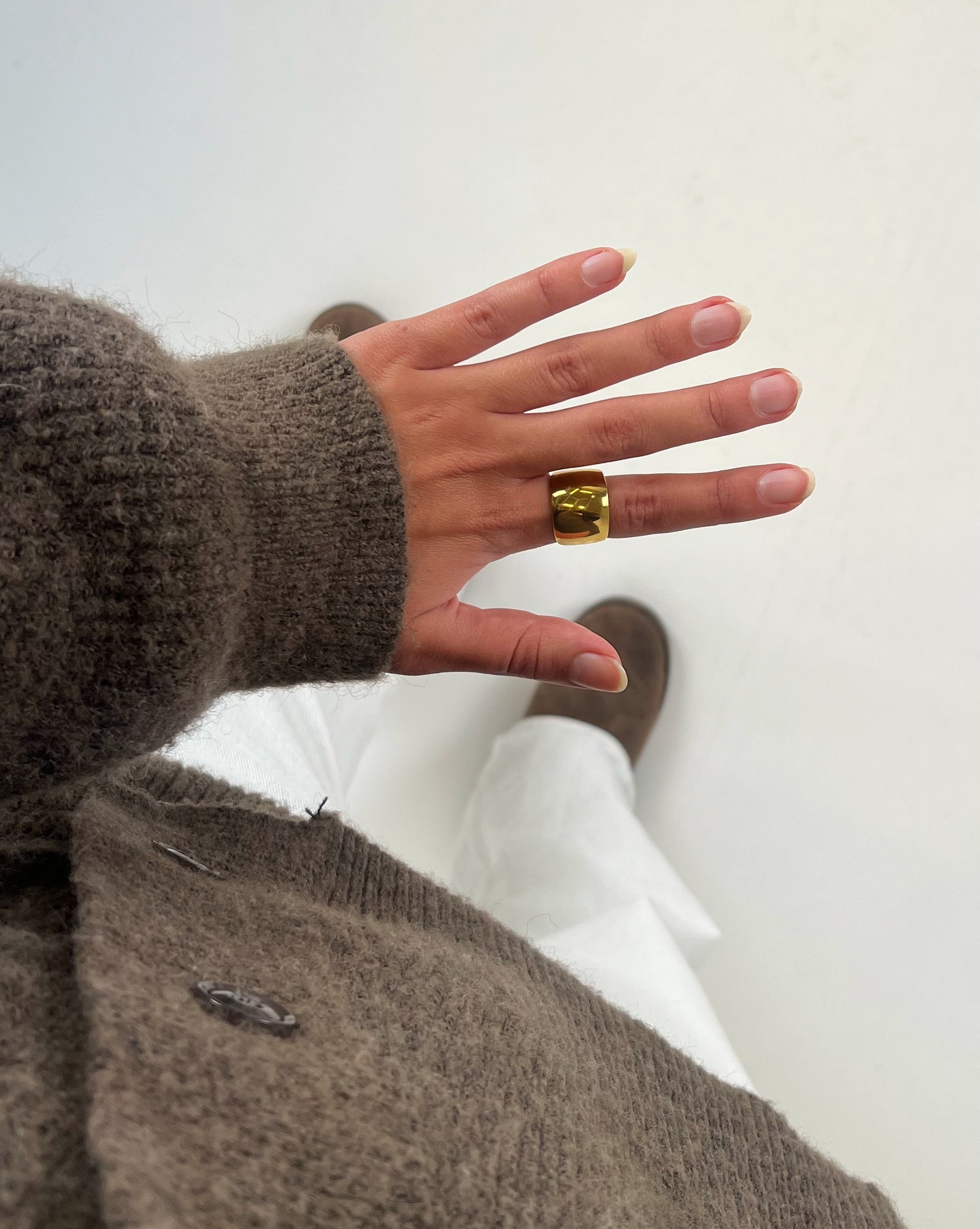 Hånd bærer en gylden ring og peger nedad. Personen har en brunt sweater og hvide bukser på. Støvler er synlige i baggrunden på en hvid baggrund.