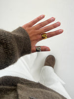En hånd med en brun sweater bærer to store ringe, én guld og én sølv. Baggrunden viser hvide bukser og en brun sko på et lyst gulv.