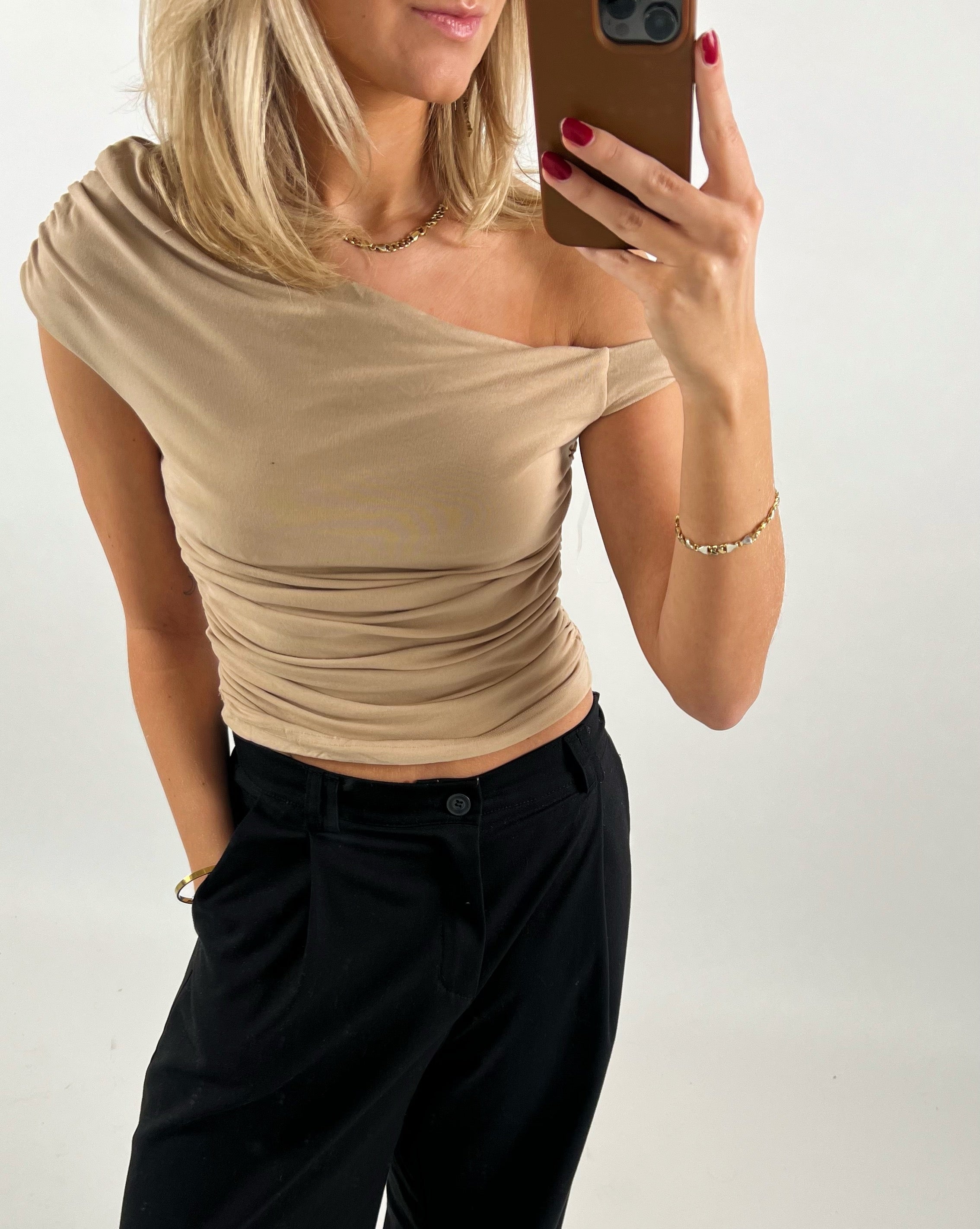 En kvinde tager en selfie iført en beige skulderbluse og sorte bukser, med rødmalede negle og guldsmykker, mod en neutral baggrund.