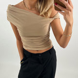 En person tager en selfie iført en beige, off-shoulder top og sorte bukser, med rød neglelak, og accessorier som øreringe og halskæde; baggrunden er neutral.