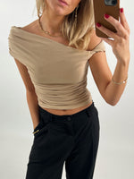 En person tager en selfie iført en beige, off-shoulder top og sorte bukser, med rød neglelak, og accessorier som øreringe og halskæde; baggrunden er neutral.