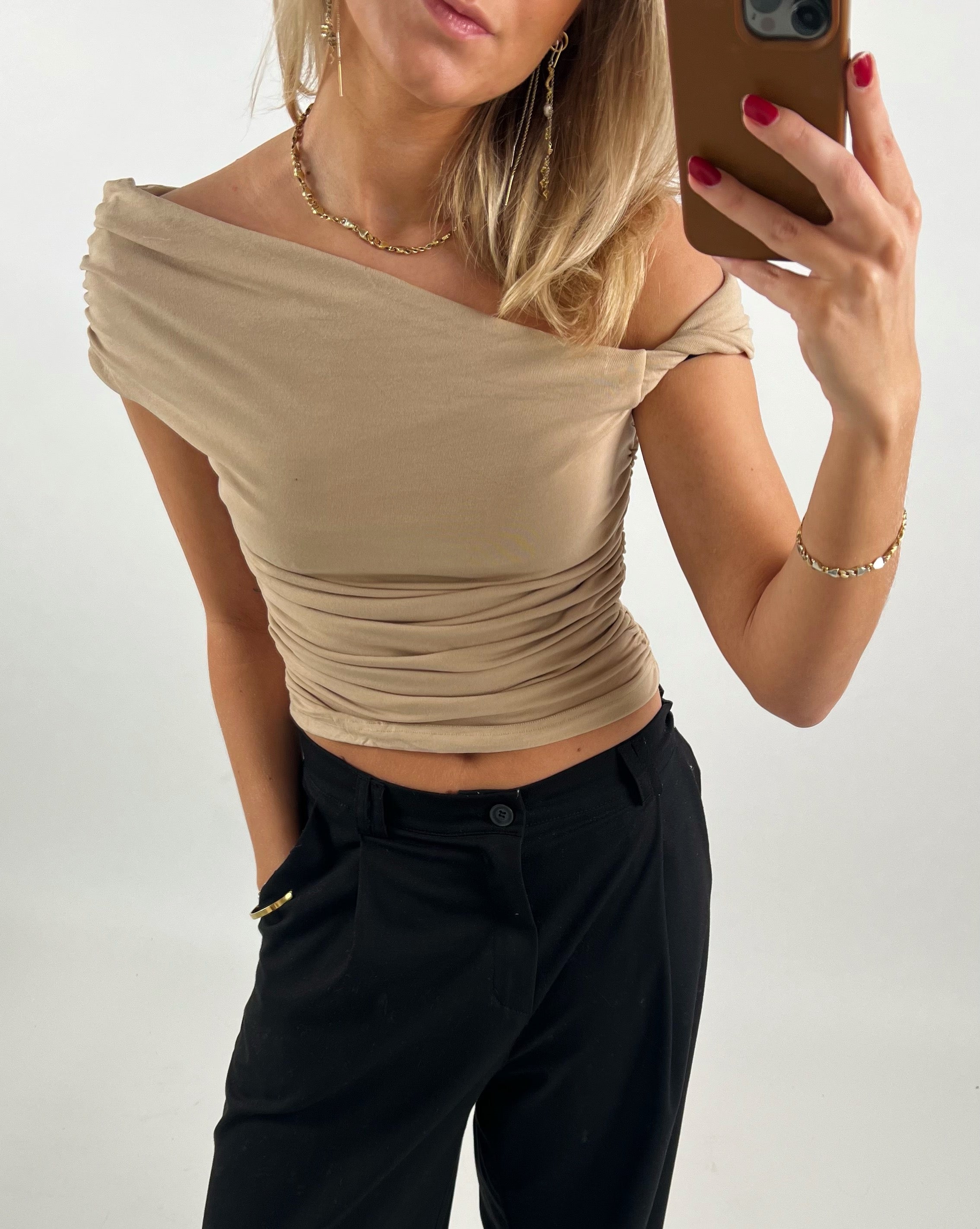 En person tager en selfie iført en beige, off-shoulder top og sorte bukser, med rød neglelak, og accessorier som øreringe og halskæde; baggrunden er neutral.