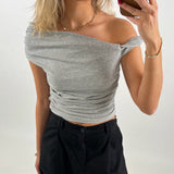 Person tager en selfie iført en grå off-shoulder top og sorte bukser. Personen har blondt hår, bærer smykker og har rød neglelak. Baggrunden er enkel og neutral.