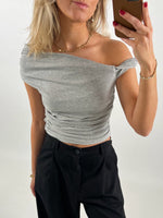 Person tager en selfie iført en grå off-shoulder top og sorte bukser. Personen har blondt hår, bærer smykker og har rød neglelak. Baggrunden er enkel og neutral.