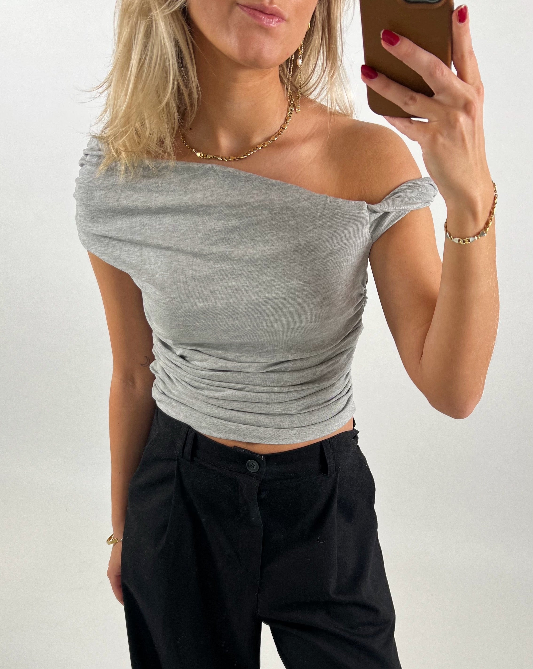 Person tager en selfie iført en grå off-shoulder top og sorte bukser. Personen har blondt hår, bærer smykker og har rød neglelak. Baggrunden er enkel og neutral.