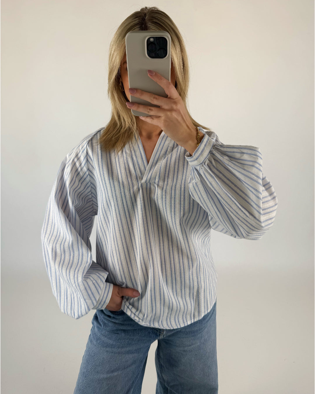 En person i en stribet skjorte tager et selfie, mens personen står mod en hvid væg. Personen holder en mobiltelefon foran ansigtet.