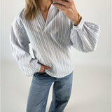 Kvinde iført stribet bluse tager selfie med telefon, står afslappet med en hånd i lommen i lyseblå jeans mod en hvid baggrund.
