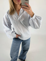 Kvinde iført stribet bluse tager selfie med telefon, står afslappet med en hånd i lommen i lyseblå jeans mod en hvid baggrund.