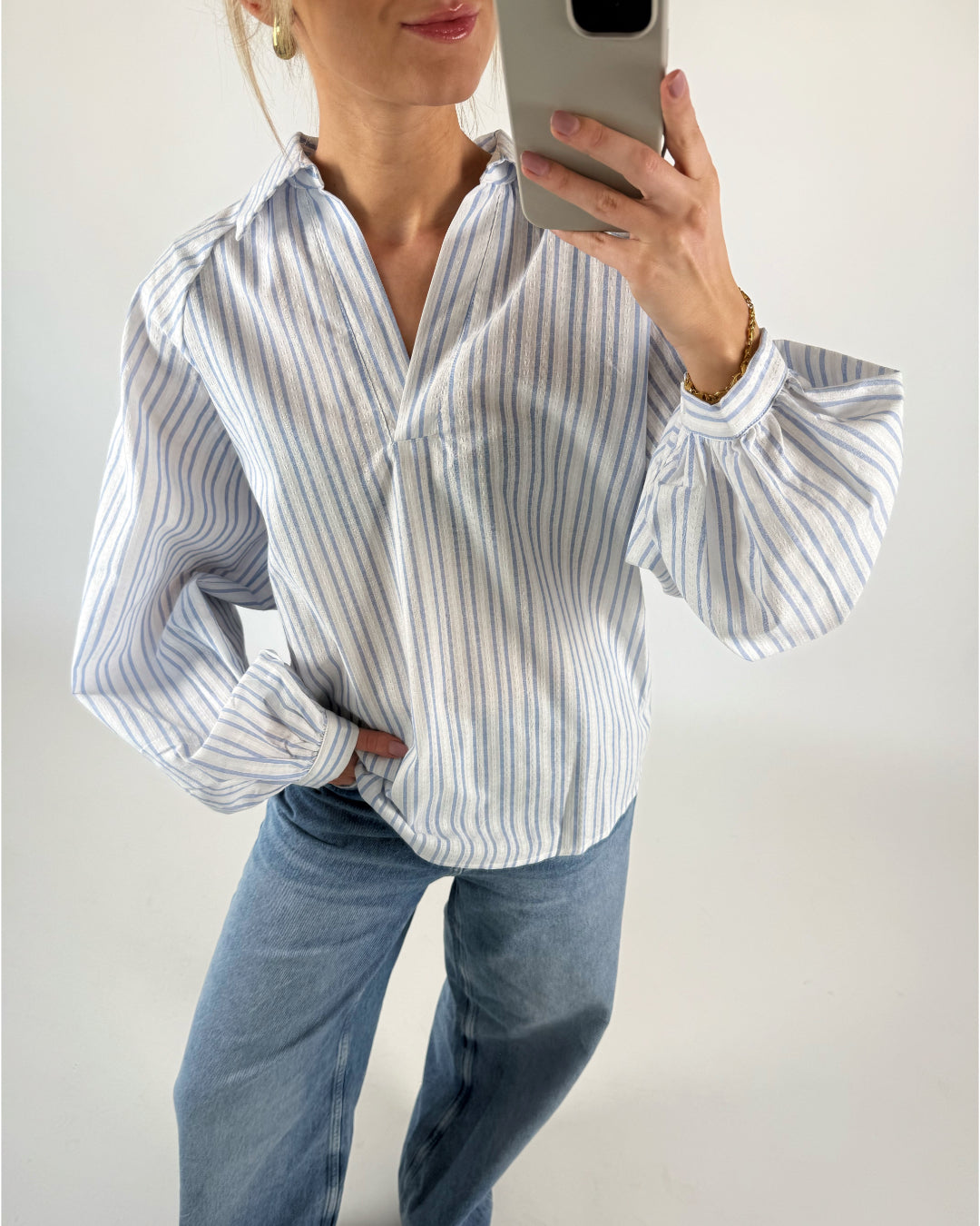 En person holder en telefon foran et spejl iført en stribet skjorte og jeans i et lyst rum.