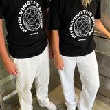 To personer iført sorte T-shirts med teksten "INVOLVING THE ENTIRE WORLD" og et cirkulært design. De står i en elevator med hvidt gulv og iført hvide bukser og sneakers.